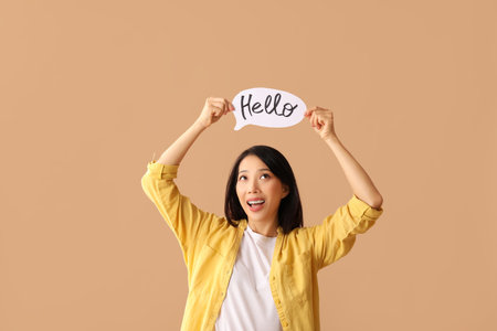 Beautiful young Asian woman holding paper speech bubble with word HELLO on beige backgroundの写真素材