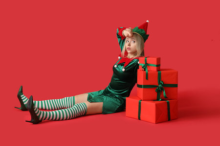 Tired woman in elf costume with Christmas gift boxes on red background