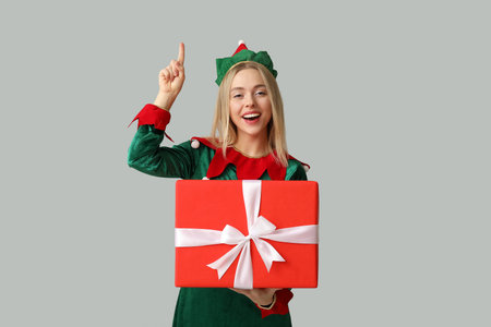 Happy young woman in elf costume with Christmas gift box pointing at something on white background