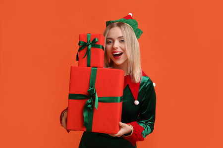 Young woman in elf costume holding Christmas gift boxes on orange background