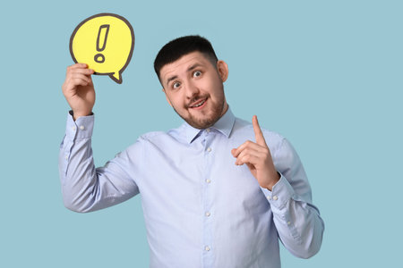Young businessman holding speech bubble with exclamation mark and pointing at something on blue...