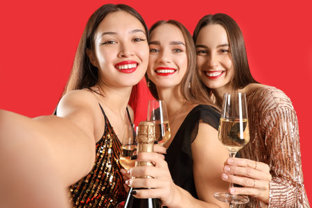 Beautiful young women with champagne taking selfie on red background, closeup. New Year celebrationの写真素材