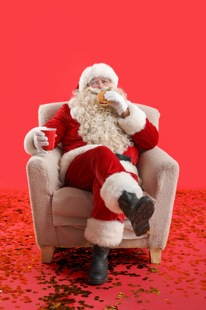 Santa Claus with cup of tea and tasty burger in armchair on red backgroundの写真素材