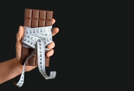 Female hand with chocolate bar and tape measure on black backgroundの写真素材