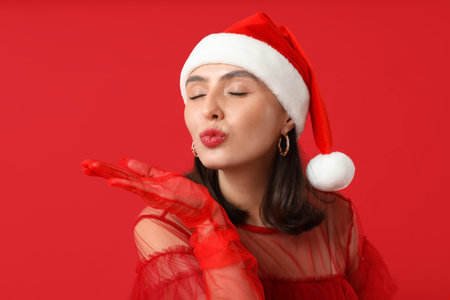 Young woman in Santa hat blowing kiss on red backgroundの写真素材