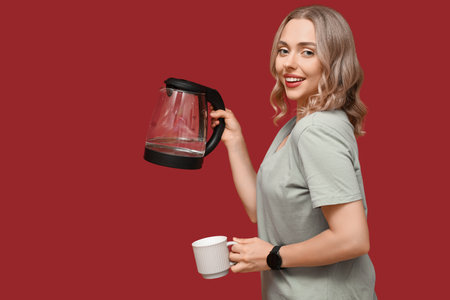 Beautiful young woman with modern electric kettle and cup on red backgroundの写真素材