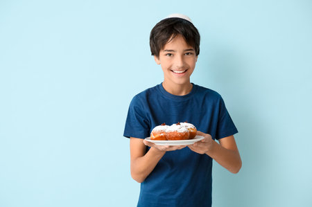 Little boy in kipa holding plate with tasty donuts on blue background. Hanukkah celebrationの写真素材