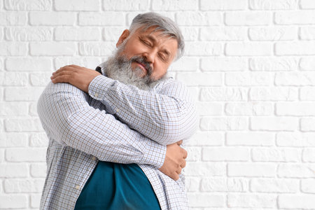 Mature man hugging himself on white brick backgroundの写真素材