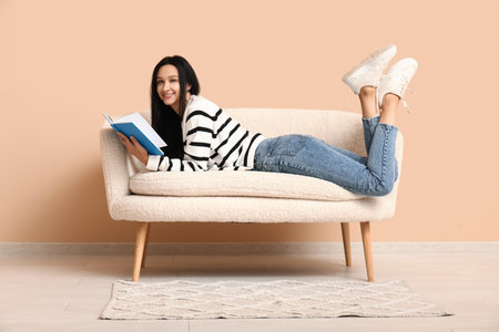 Beautiful young woman resting on comfortable sofa near beige wall and reading bookの写真素材
