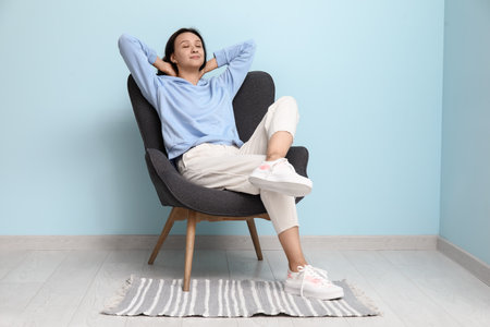 Beautiful young woman resting in comfortable armchair near blue wallの写真素材