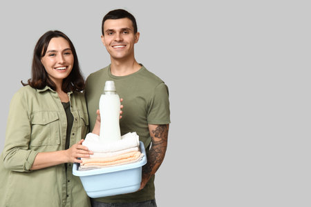 Young couple with laundry basket and bottle of detergent on gray backgroundの写真素材