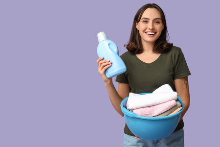 Young woman with laundry basket and bottle of detergent on lilac backgroundの写真素材