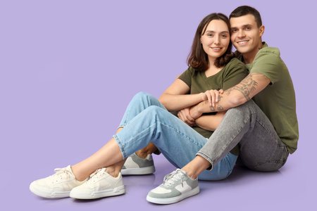 Happy young couple sitting on floor and hugging against lilac backgroundの写真素材