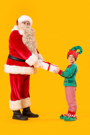 Santa Claus and cute little elf with Christmas gift on yellow backgroundの写真素材