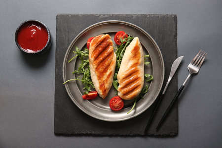 Plate of tasty grilled chicken breast with arugula, tomatoes and ketchup on gray backgroundの写真素材