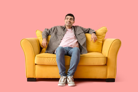 Handsome young man sitting on yellow sofa against pink backgroundの写真素材