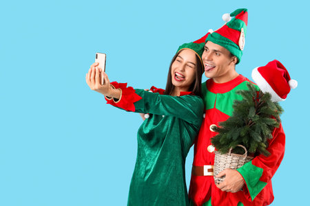 Happy couple in elf's costumes with Christmas tree and mobile phone taking selfie on blue background