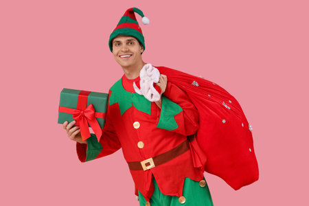 Handsome young man in elf's costume with Santa bag and gift box on pink backgroundの写真素材