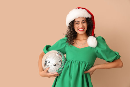 Beautiful African-American woman in Santa hat with disco ball on beige background. New Year celebrationの写真素材