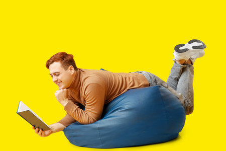 Young man lying on beanbag and reading book on yellow backgroundの写真素材