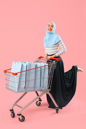Beautiful Muslim woman with trolley and shopping cart on pink backgroundの写真素材