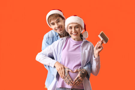 Young pregnant couple in Santa hats with paint brush hugging on orange background