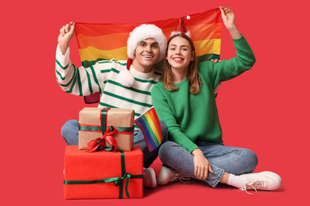 Young couple in Santa hats with LGBT flag and Christmas gifts sitting on red backgroundの写真素材