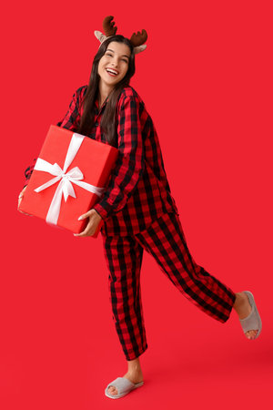 Happy young woman in checkered pajamas and reindeer horns with Christmas gift box on red backgroundの写真素材