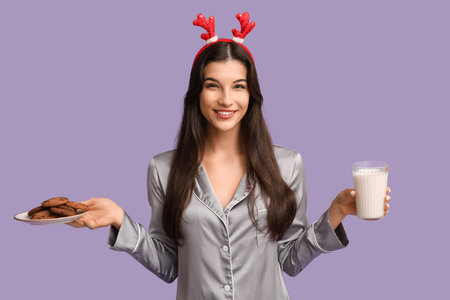 Beautiful young woman in gray pajamas with sweet cookies and glass of milk on purple backgroundの写真素材
