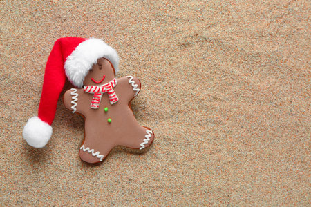 Tasty gingerbread cookie with Santa hat on sand background