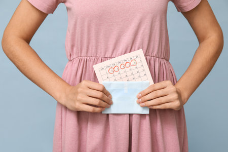 Beautiful young woman holding pad and calendar with marked days of menstruation on blue backgroundの写真素材