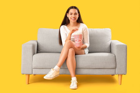 Young woman with popcorn watching movie on sofa against yellow backgroundの写真素材