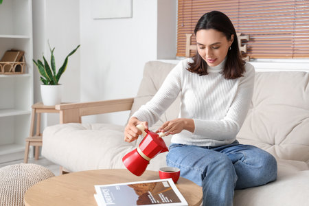 Pretty young woman with geyser coffee maker and cup of espresso sitting on sofa in living roomの写真素材