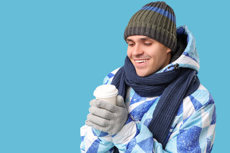 Young man in stylish puffer jacket and scarf with paper cup of coffee on blue backgroundの写真素材