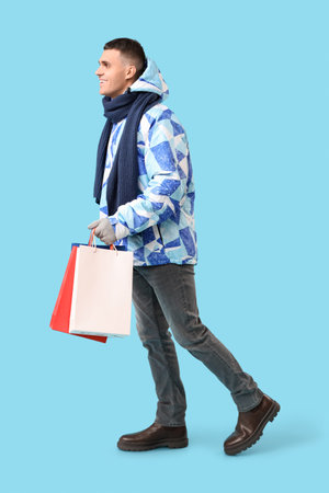 Young man in stylish puffer jacket and scarf with shopping bags on blue backgroundの写真素材