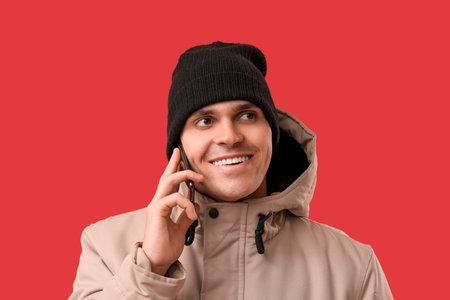 Young man in stylish puffer jacket and hat talking by mobile phone on red background