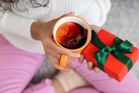 Young African-American woman with cup of hot mulled wine and gift box on carpet at homeの写真素材