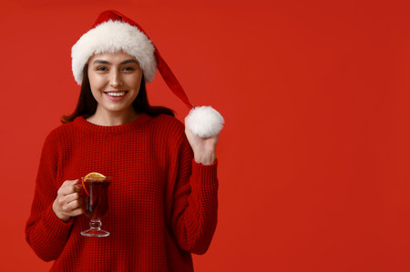 Beautiful young woman in Santa hat with glass cup of hot mulled wine on red backgroundの写真素材