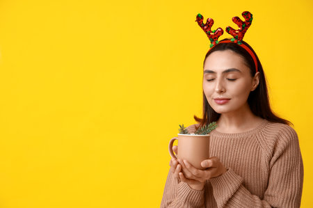Beautiful young woman in reindeer horns with cup of hot mulled wine on yellow backgroundの写真素材
