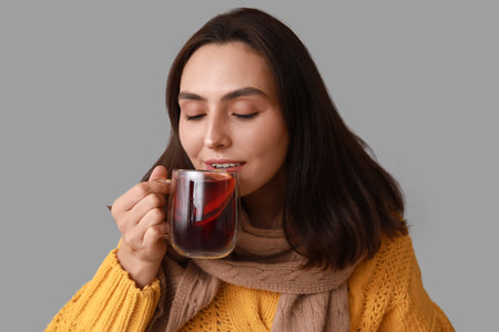 Beautiful young woman in scarf with glass cup of hot mulled wine on gray backgroundの写真素材
