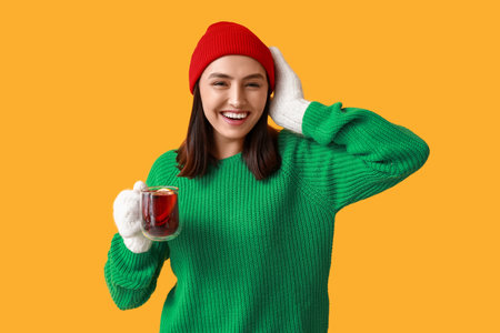 Happy young woman with glass cup of hot mulled wine on yellow backgroundの写真素材