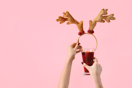 Female hands with glass of warm mulled wine and Christmas reindeer horns on pink backgroundの写真素材