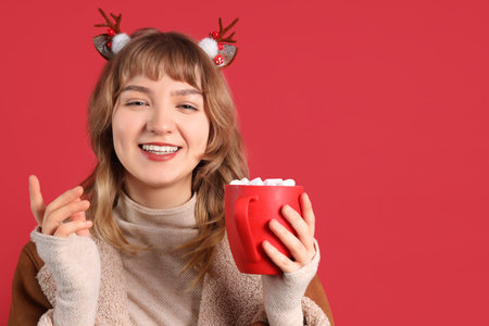 Pretty young woman holding cup of hot chocolate with marshmallows on red backgroundの写真素材