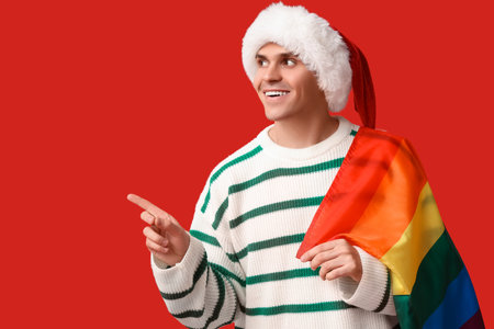 Young man in Santa hat with LGBT flag pointing at something on red backgroundの写真素材