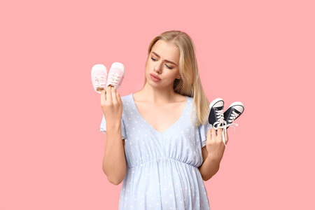 Beautiful thoughtful pregnant woman with baby shoes on pink background