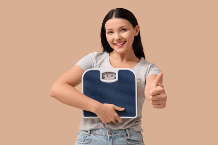 Beautiful young woman with scales showing thumb-up gesture on beige background. Weight loss conceptの写真素材