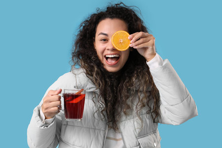 Beautiful young African-American woman with glass cup of hot mulled wine and orange on blue...