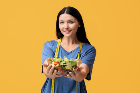 Beautiful young happy woman with measuring tape and bowl of fresh salad on yellow background. Weight loss conceptの写真素材