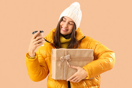 Young woman in winter clothes with Christmas gift and coffee cup on beige backgroundの写真素材