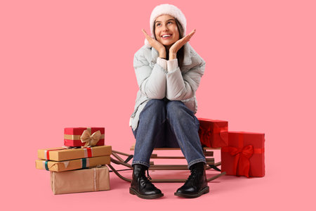 Young woman in Santa hat with Christmas gifts sitting on sledge against pink background